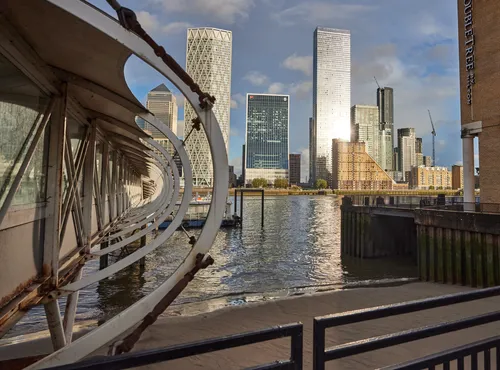 This urban architectural photograph shows the Canary Wharf hotel pier in London, United Kingdom, during a late afternoon in early autumn. The pier’s curved structure frames the scene, while the River Thames flows alongside it, reflecting light from the tall buildings of Canary Wharf in the background. The iconic skyscrapers, such as One Canada Square and other structures of the Canary Wharf financial district, are visible and create a striking cityscape. The photograph captures the urban atmosphere of London, highlighting the blend of contemporary architecture with the rippling water of the River Thames under autumn sunlight.